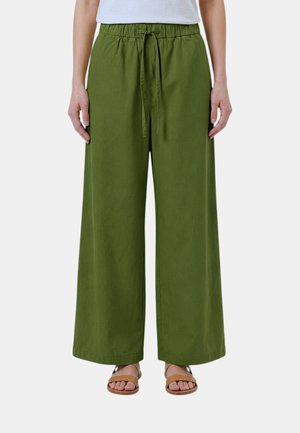 Persona che indossa pantaloni verdi a gamba larga con vita regolabile a coulisse e sandali marroni con punta aperta, in piedi davanti a uno sfondo semplice.