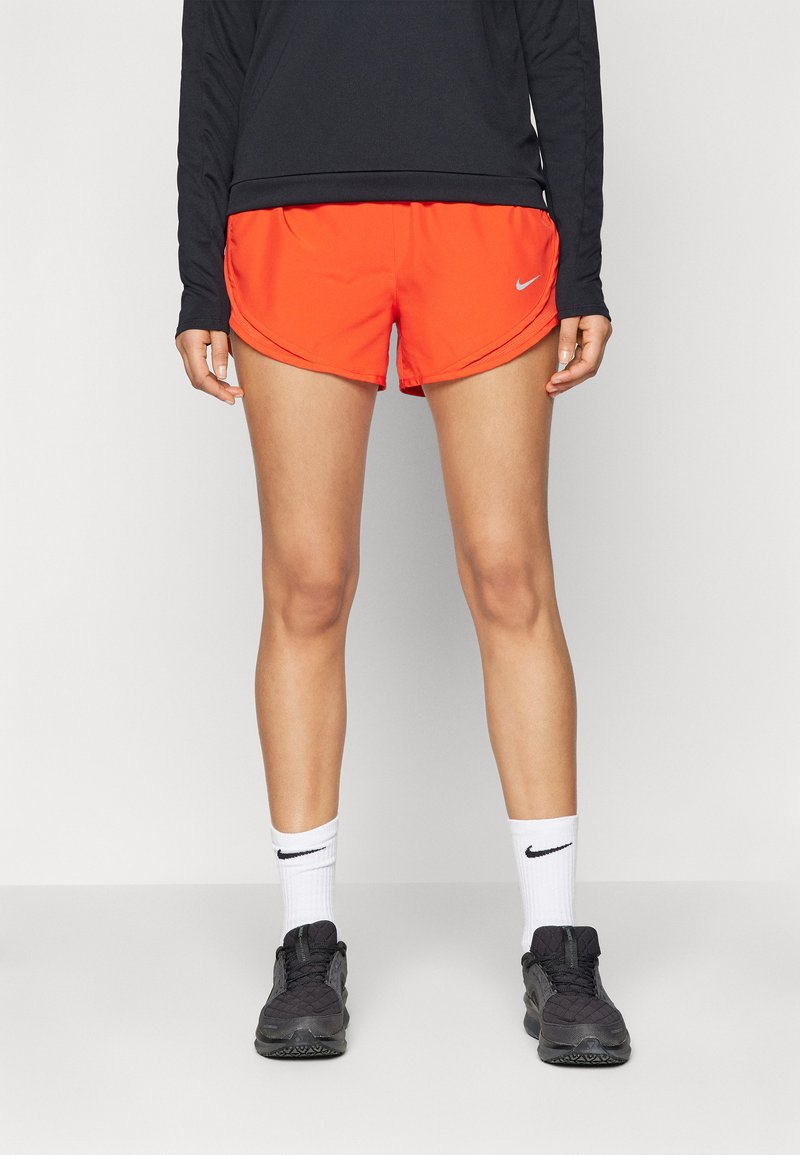 Shorts de course pour femmes de Nike, orange vif, en tissu léger, hem incurvé, associés à des chaussures de sport noires et des chaussettes blanches Nike.