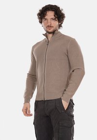 Maglione zip-up a costine marrone chiaro con maniche lunghe, colletto alto e cerniera a contrasto. Il tessuto a maglia testurizzato aggiunge interesse visivo.