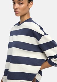 Sweter w biało-niebieskie paski z okrągłym dekoltem, luźnym krojem i miękką fakturą; posiada długie, lekko bufiaste rękawy.