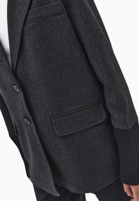 Tummanharmaa bleiseri, jossa on napit, läppätaskut ja ribbimuotoillut mustat hihnat, käytetty valkoisen paidan ja tummien housujen päällä.