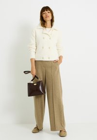 Cárdigan de punto doble botonadura en crema, pantalones anchos beige, bolso marrón con detalles de tachuelas y zapatos sin cordones con estampado. Fondo liso.