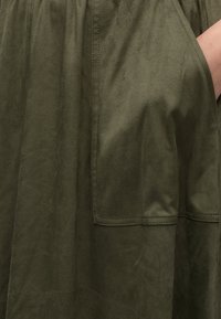 Main dans la poche d'un vêtement en daim vert olive montrant la texture et les détails de couture sur un tissu doux.