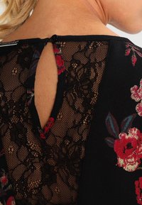Top de encaje negro con bordado floral en tonos rojos y azules, con una apertura en forma de llave en la parte posterior y cierre de botón.