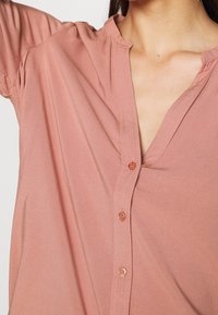 Blouse rose clair avec un décolleté en V, des manches courtes et une fermeture à boutons. Texture lisse avec un subtil détail de couture le long du décolleté.