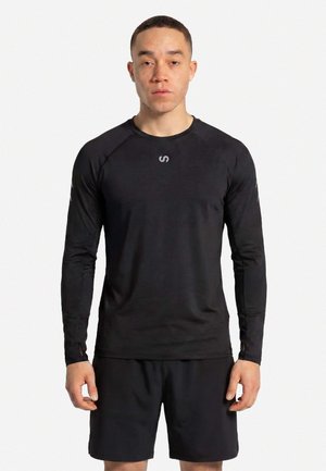 Uomo in piedi di fronte, indossa una maglia sportiva a maniche lunghe nera con fori per i pollici e pantaloncini neri su sfondo bianco.