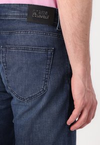 Tamno plavi traperice s leđnim džepom i crnom kožnom etiketom s utisnutim tekstom. Glatka tekstura i ravna silueta.