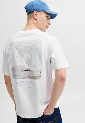 Young man wearing a blue cap and white T-shirt with a coastal scene and text "Plage de la Douane Beach & Social Club" on the back.