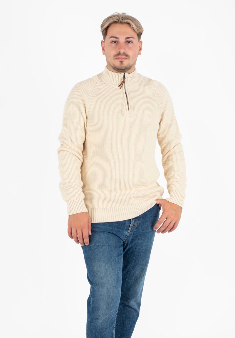Beżowy sweter prążkowany z wysokim kołnierzem i półzamkiem. Noszony na niebieskich dżinsach, charakteryzujący się luźnym krojem i fakturowanym splotem.