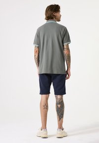 Grijze polo met kraag, marineblauwe shorts en lichte sneakers. Heeft gestreepte accenten op de kraag en gedetailleerde been tattoos.