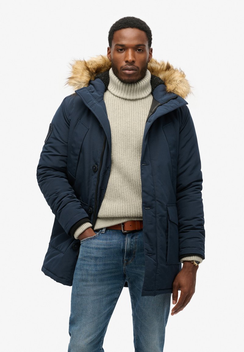 Cappotto puffer blu con cappuccio foderato in pelliccia sintetica, chiusura frontale con bottoni, due tasche laterali, indossato sopra un maglione a collo alto color crema e jeans blu.