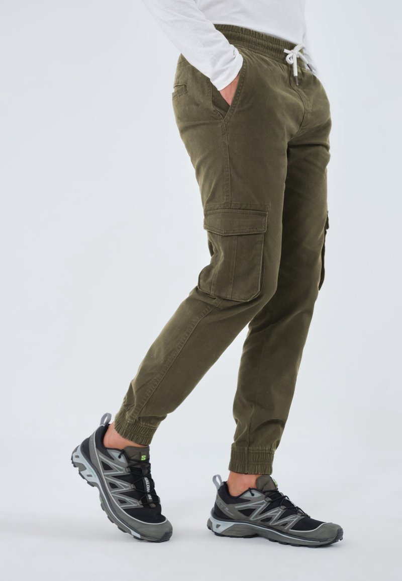 Pantalon cargo vert olive avec taille élastique, poches latérales et chevilles fuselées, associé à des chaussures de sport noires et grises avec une semelle texturée.