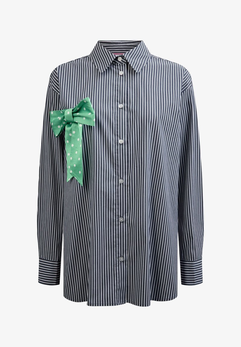 Chemise à manches longues boutonnée aux rayures verticales noires et blanches avec un grand nœud à pois verts fixé sur la partie gauche de la poitrine.