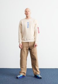 Μπεζ φούτερ με μπλε και κόκκινες ετικέτες, καμηλό παντελόνι cargo και μπλε-μαύρα αθλητικά παπούτσια με σχέδια, που στέκεται σε μπλε χαλί.