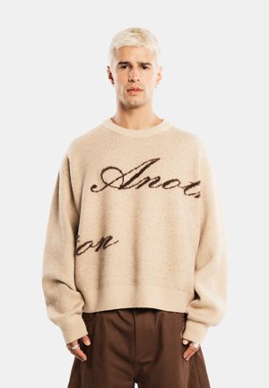 Ung person med kort blondt hår iført en beige sweater med mørk skrift og løse brune bukser, der står mod en ensfarvet baggrund.