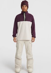 Purple and white pullover with half-zip and logo on collar, paired with light-colored cargo pants and white boots. Soft fabric, relaxed fit.
