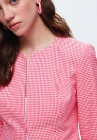 Blazer a scacchi rosa con scollo rotondo e chiusura frontale singola. Presenta un tessuto liscio e una vestibilità strutturata, abbellito da orecchini rosa.
