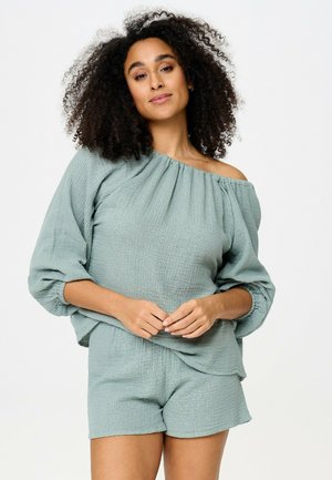 Frau mit lockigem Haar, die ein locker sitzendes, hellgrünes schulterfreies Oberteil und passende Shorts trägt und mit vorgehaltenden Händen dasteht.