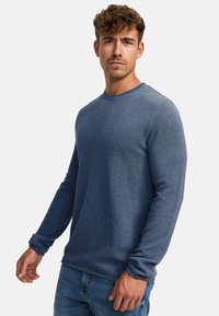 Sininen pitkähihaisen teksturoitu neulepaita pyöreällä kaula-aukolla, valmistettu neulottusta materiaalista. Yhdistetty farkkuihin, kädet rentoutuneet sivuilla.