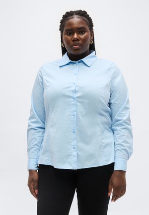 Γυναίκα με πλεξίδες φοράει ανοιχτόγαλανό πουκάμισο με κουμπιά και μαύρο παντελόνι, κοιτάζοντας μπροστά με ουδέτερη έκφραση σε απλό φόντο.