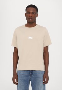 Beige bomulds t-shirt med korte ærmer og rund hals, med et kontrastfuldt hvidt MK-logo på brystet, parret med blå jeans.