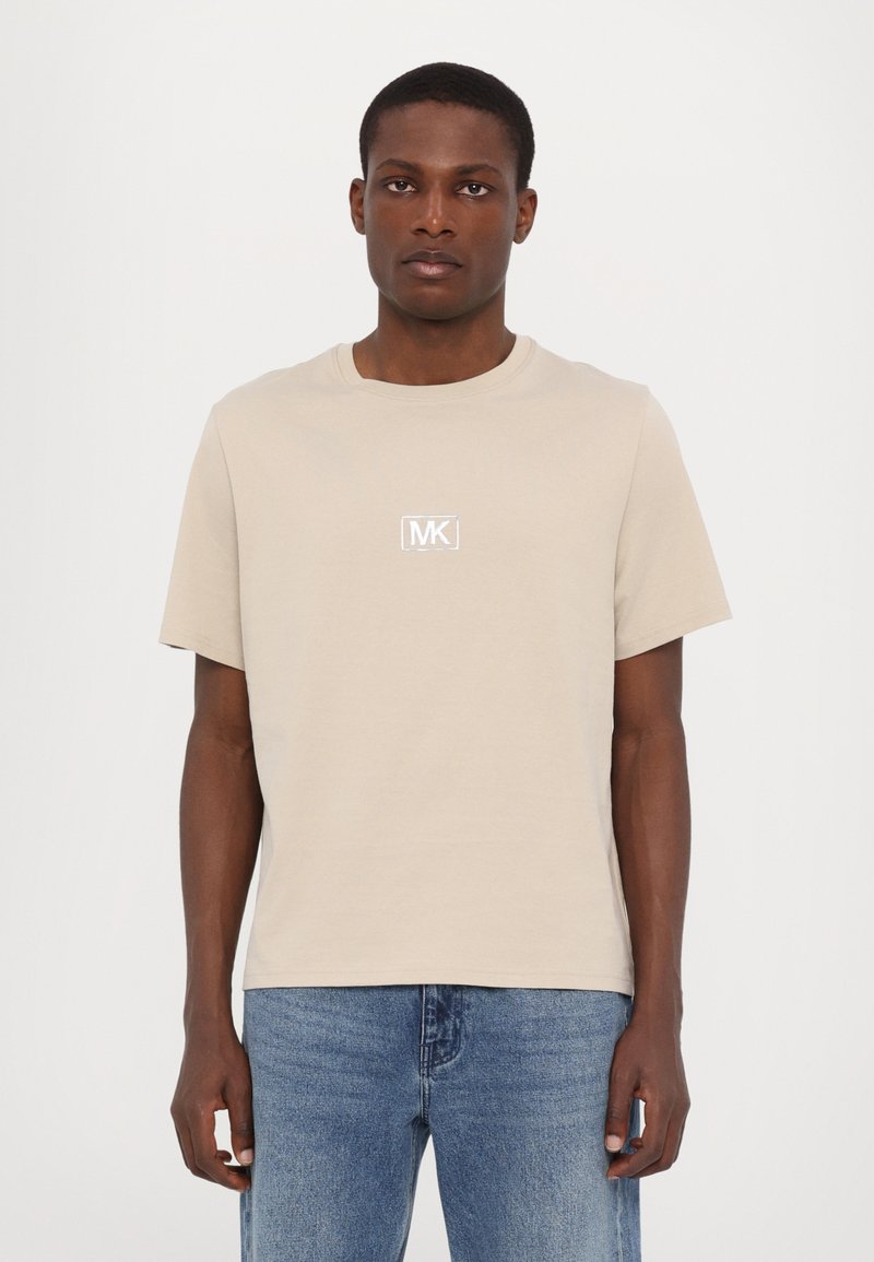 Beige bomulds t-shirt med korte ærmer og rund hals, med et kontrastfuldt hvidt MK-logo på brystet, parret med blå jeans.