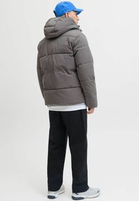 Mężczyzna zwrócony bokiem, ubrany w szarą kurtkę puchową z kapturem, niebieską czapkę z daszkiem, czarne spodnie i białe trampki na jednolitym tle.