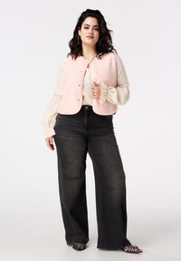 Lichtroze, gestructureerd mouwloos vest met knoopsluiting, gedragen over een crème blouse. Gecombineerd met high-waist donkerblauwe spijkerbroek en gebloemde schoenen.