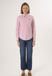 Chemise longue à rayures rouges et blanches avec un col boutonné et un logo en forme de cœur, assortie à un jean bleu à jambe droite et des chaussures brunes.