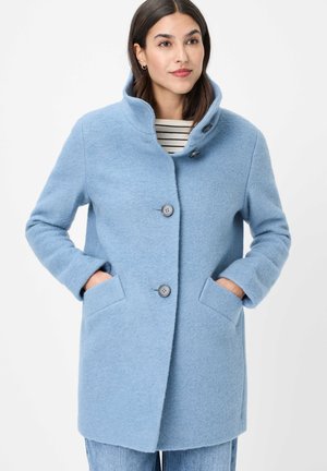 Frau trägt einen hellblauen Wollmantel mit großen Knöpfen und Kragen, die Hände in den Taschen, und darunter ein gestreiftes Hemd.