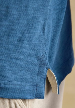 Sinine kudumiskangast särgi äär küljesõlmega, alt paistavad beežid tekstuuriga püksid.