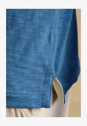 Mėlynos megztos medžiagos marškinių apačia su šoniniu plyšiu, pro kurį matomos smėlio spalvos tekstūruotos kelnės.