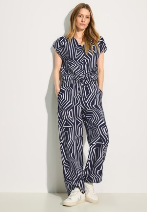Marineblauer Jumpsuit mit weißem geometrischem Muster, kurzen Ärmel, elastischem Bund und Seitentaschen; kombiniert mit weißen Sneakers.