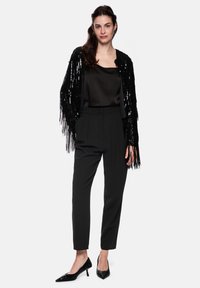 Schwarze Jacke mit Paillettenfransen über einem schwarzen Satintop, kombiniert mit hoch taillierter schwarzer Hose und spitzen schwarzen High-Heels.