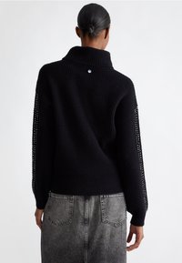 Maglione nero lavorato a maglia con colletto alto, caratterizzato da dettagli a rete lungo le maniche. Abbinato a jeans grigi a vita alta.