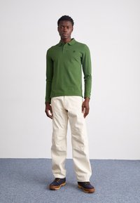 Polo verde de manga longa com um pequeno logótipo, combinado com calças cargo bege e sapatos escuros com solas amarelas sobre um fundo claro.