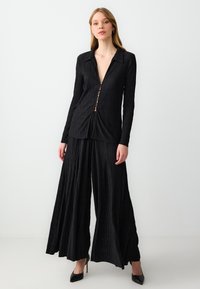 Zwart geplooid set bestaande uit een blouse met knopen en een diepe halslijn, en een wijde broek. Gladde stof met een gestructureerd oppervlak. Hoge hakken.