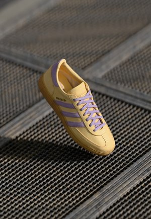 Żółty sneaker Adidas Spezial z fioletowymi paskami i sznurowadłami, unoszący się nad teksturowanym metalowym rusztem w tle.