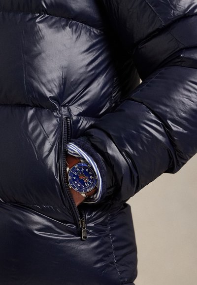 Arm in a shiny navy puffer jacket pocket, showing a wristwatch with a blue face and brown leather strap over a striped shirt cuff.