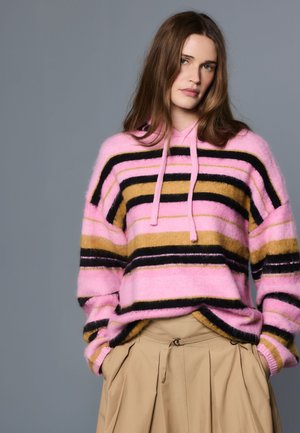 Femme aux cheveux bruns portant un sweat à capuche rayé rose, noir et moutarde et un pantalon plissé beige, debout devant un fond gris.