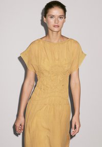 Fließendes, senfgelbes Kleid mit gerafftem Oberteil, kurzen, flatterhaften Ärmeln und strukturierten Details entlang der Taille und des Saums.