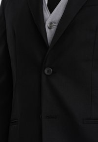 Mustassa pikkutakissa on sileä pinta, yksittäinen nappi ja hillitty taskutyyli. Se on kerroksittain vaaleanharmaan liivin päällä.