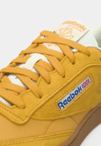 Kratki prikaz senf žutih Reebok Classic tenisica s platnenim vezicama i plavim logotipom s britanskom zastavom na bočnoj strani.