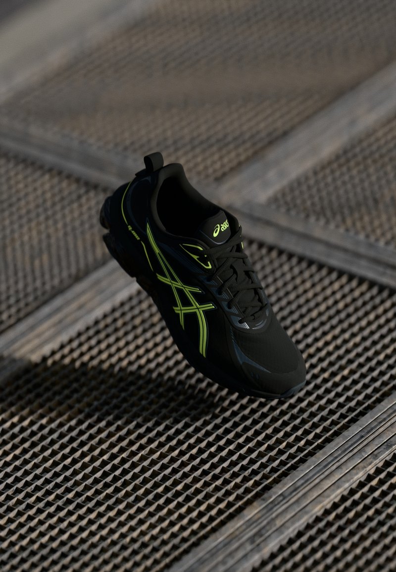 Chaussure de course Asics noire et vert néon avec lacets sur une surface en grille métallique texturée en faible lumière.
