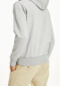 Osoba ubrana w jasnoszary hoodie i beżowe spodnie, z rękami w kieszeniach, odwrócona plecami do aparatu na białym tle.