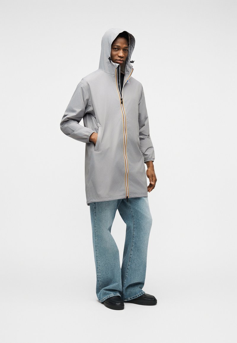 Jeune personne portant un imperméable à capuche gris avec une fermeture éclair rayée, un jean bleu et des chaussures noires, debout avec une main dans la poche.
