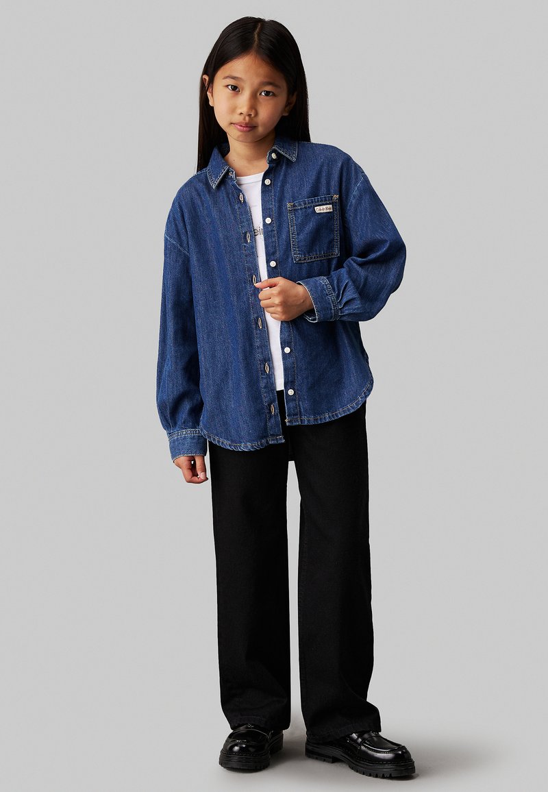 Camicia di jeans blu scuro con maniche lunghe, chiusura a bottoni, una tasca sul petto e una vestibilità comoda, abbinata a pantaloni a gamba larga neri e scarpe lucide.