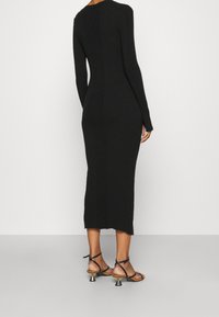 Robe noire en maille côtelée avec manches longues, coupe ajustée et longueur mi-mollet. Dotée d'une fente à l'arrière. Associée à des sandales noires à talons avec brides.