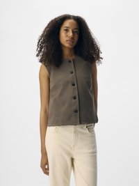Haut sans manches brun avec un devant à boutons, fabriqué en tissu lisse, associé à un jean couleur crème, se tenant devant un fond neutre.