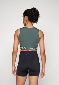 Zeleni prirezani top s trakom "NIKE PRO" na pasu, v kombinaciji s črnimi kratkimi hlačami visoko pasu, ki na zadnji strani nosijo majhen logo.